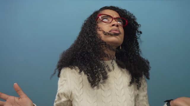 Woman with curly hair wearing red glasses and white cable knit sweater raises hands palms up in studio; confusion.
