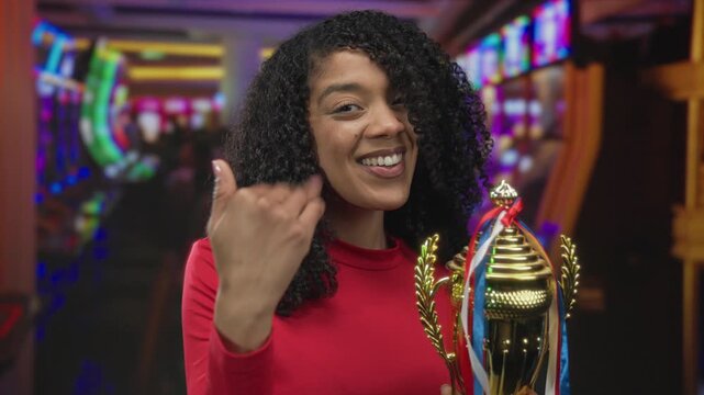 Woman holds gleaming trophy and shows thumbs up gesture in brightly lit casino slot machine hall; joy celebration success achievement.