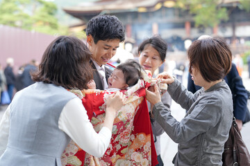 A group of people are gathered around a baby, with one woman holding the baby and another woman holding a red blanket. The scene is lively and joyful, with everyone smiling and enjoying the moment