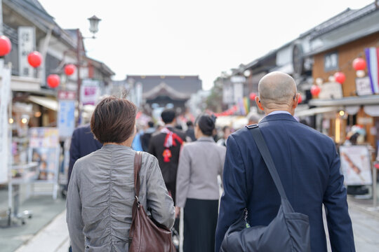 A man and woman walk down a street with many people around them. The man is wearing a suit and the woman is wearing a gray jacket. Scene is busy and bustling, with people walking and carrying bags