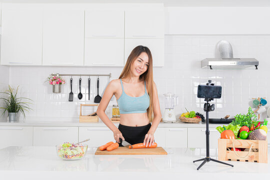 Smiling young woman in blue workout clothing chopping carrot in her modern kitchen while recording a video on her smartphone, sharing her healthy lifestyle for online.
