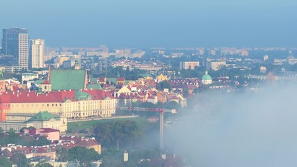 Aerial view of Warsaw’s Royal Castle and Old Town rooftops emerging above dense morning fog drifting over Vistula River, with modern city skyline in distance. Wide shot. - Powered by Adobe