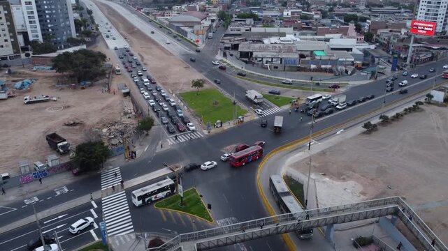A newly built extension to an express way, named "Via Expresa Sur". Cars drive on the intersection below as the drone hovers above and pans left while tilting up showing the cityscape of Lima, Peru.
