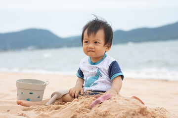 Fototapeta premium Asian family baby sitting on sandy beach with bucket and shovel smiling at seaside
