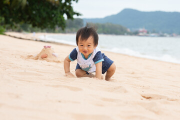 Asian happy family baby crawling on sandy beach with ocean waves and distant hills smiling with playful expression