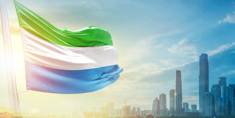Sierra Leone flag wavering in beautiful clouds with building skyline. The flag with drone shot in beautiful sky. Sierra Leone national flag for independence day.
