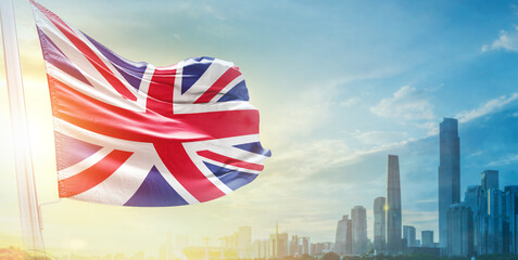 Obraz premium United Kingdom flag wavering in beautiful clouds with building skyline. The flag with drone shot in beautiful sky. UK national flag for independence day.