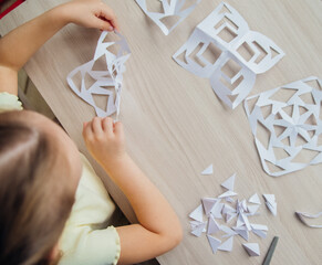 A child creating paper white snowflakes at a table at home. A child cuts patterns on paper with...