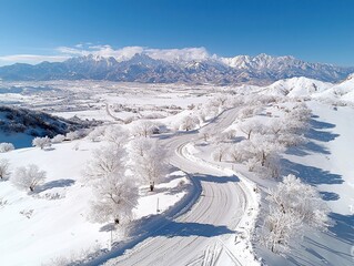 Aerial view of a snowy road winding through a winter landscape with snow-covered trees and mountains in the background under a clear blue sky. The scene evokes