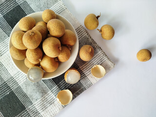 Flat lay a pile of whole longans on a white plate over a checkered cloth, with scattered and peeled fruits showing the flesh, set against a clean white background with ample copy space.