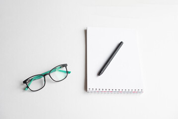 A pair of black glasses with green accents rests beside a blank notepad and a black pen on a white surface. Ideal for office or study themes.
