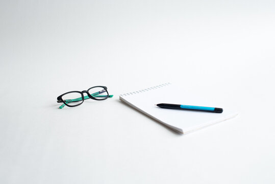 A pair of black glasses, a blue pen, and a blank notepad are placed on a white surface. The scene conveys a sense of organization and readiness for writing.