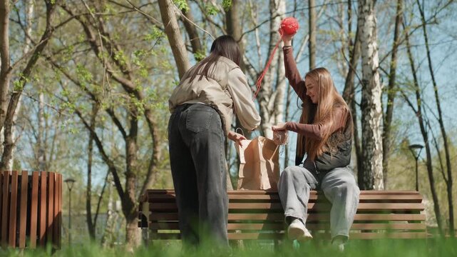 two friends caucasian and asian exchanging a red balloon and snacks on bench in sunny park, passing paper bag items while laughing and offering cups; spontaneous playful moment, spring birch grove,