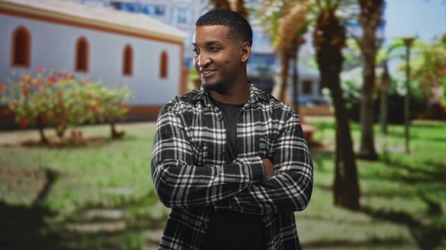 Young black man in plaid shirt with arms crossed and a soft smile, standing near palm trees and arched windows of a building; confidence.