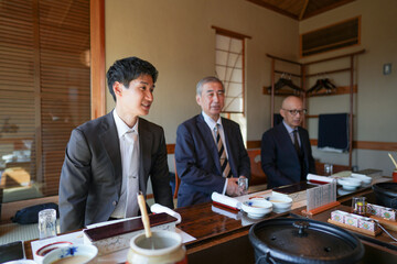 Three men are sitting at a table in a room, dressed in suits and ties. One of the men is smiling, while the other two look serious. The room has a traditional Japanese design, with wooden furniture