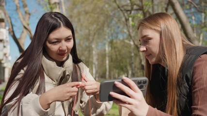 friends inspect small gadget beside phone, asian and white women examine earbuds packaging with careful hands, closeup of fingers and device, curious expressions and unboxing vibe in sunny park