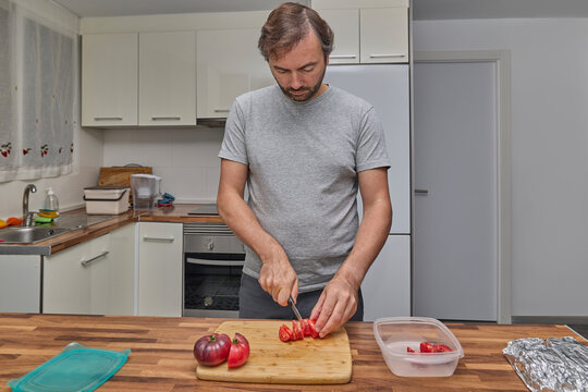 A man cuts ripe tomatoes on a wooden board while preparing fresh ingredients in the kitchen.