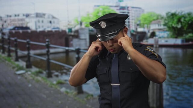 Uniformed policeman covers ears using fingers to block water canal noise on a bustling city street walkway; discomfort.