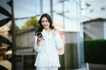 Fototapeta premium An Asian woman happily talking on the phone while holding shopping bags outside a modern building, capturing the joyful lifestyle of a young Asian woman on a shopping day.