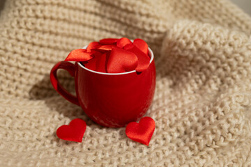 A bright red mug filled with fabric heart shapes sits on a textured, warm beige blanket. The scene captures the essence of love and warmth perfect for Valentine's Day celebrations.