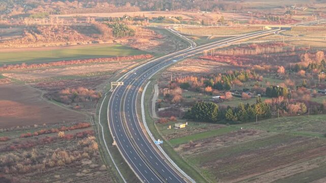 An aerial view of a winding highway cutting through expansive rural fields and scattered vegetation. Soft, warm light highlights the surrounding farmland, small houses, and patches of forest.