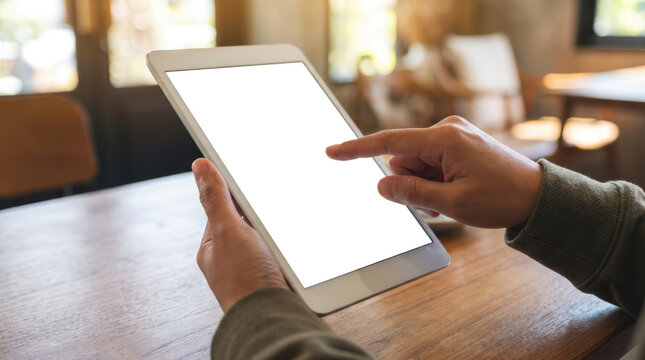 A person's hands holding a white tablet computer with a transparent screen touching the screen with a finger on a wooden table with a blurred background of a cafe interior - Powered by Adobe