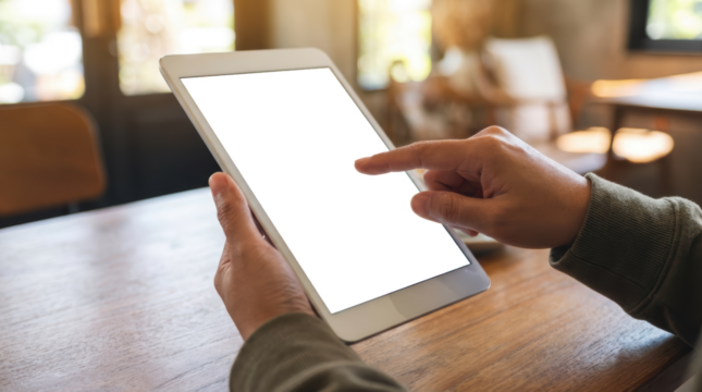 A person's hands holding a white tablet computer with a transparent screen touching the screen with a finger on a wooden table with a blurred background of a cafe interior