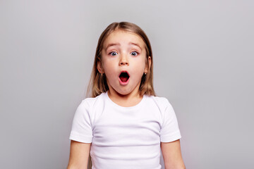 Surprised young girl with wide open mouth looking at camera in studio, wearing white t-shirt, expressive emotional portrait against clean grey background