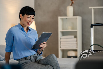 Asian business woman in a modern hotel room using a tablet beside her luggage, making online bookings and checking travel options for her business trip.