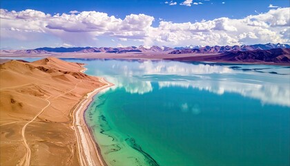 An aerial view of a turquoise lake with mountains in the background under a partly cloudy sky. The landscape features a sandy shoreline and the reflection of th