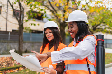 Two women engineers wearing hard hats and safety vests discussing a blueprint outdoors,...