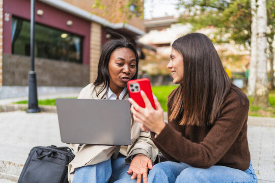 Two young diverse women students sitting outdoors, engaging in a discussion and sharing content on a laptop and smartphone
