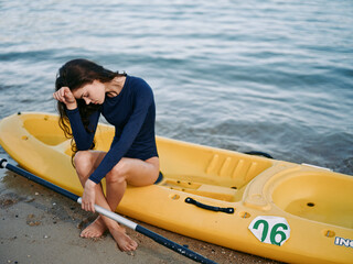A young woman sits in a yellow kayak on a sandy shore, leaning forward with an elbow on her knee and a hand resting on her brow, lost in quiet reflection.