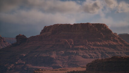 Red rock formations rise dramatically against a backdrop of clouds in the desert. The warm light of sunset casts beautiful shadows, highlighting the rugged terrain and natural beauty.