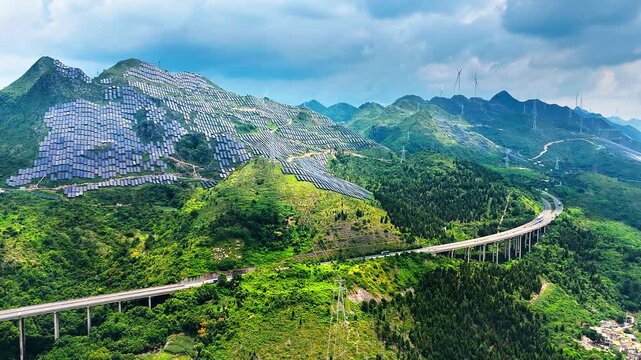 Aerial shot of solar panels covering green mountain slopes with highway bridge and wind turbines under cloudy sky