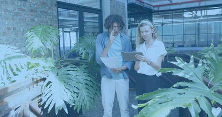 Discussing two colleagues wearing office wear, holding tablet, papers and phone in atrium with pots