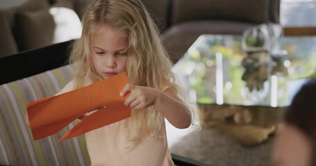 Inspecting orange paper with cuts, girl wearing pale pink top sitting on striped cushion