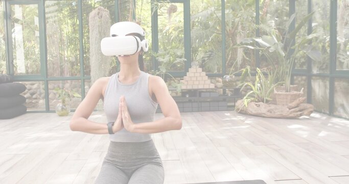 Kneeling midadult woman meditating on mat in glass studio with plants, wearing grey leggings, VR