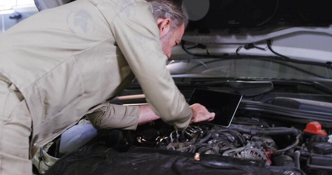 Leaning senior mechanic in beige coveralls inspecting engine at garage, using tablet  and  fender co