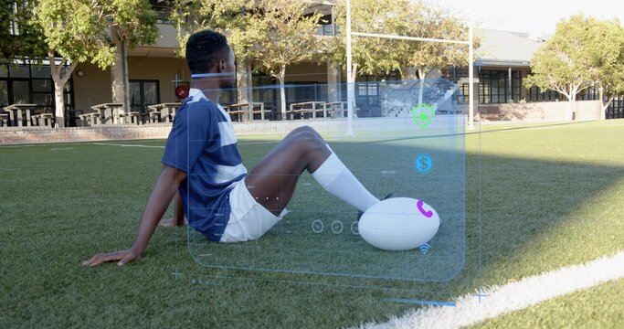 Sitting athlete in sportswear on campus turf, pressing foot on white oval ball, viewing digital UI