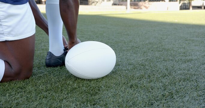 Kneeling player long-white-socks tying black cleat touching white-oval ball on turf, copy space