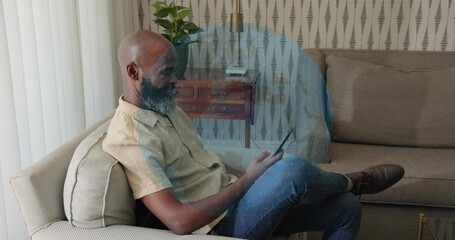 Sitting man holding tablet on beige sofa at home, wearing light short-sleeve shirt and blue jeans
