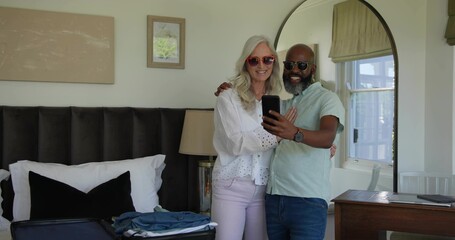 Taking selfie senior couple wearing shades, smiling and posing in bedroom with phone, open suitcase