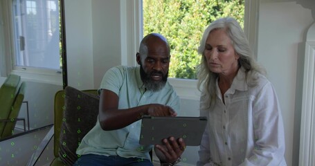 Pointing at tablet, mature pair leaning on sofa by window, with patterned cushion and green chair