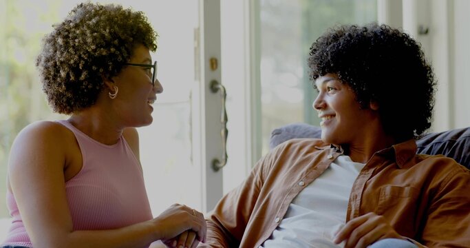 Talking couple on couch exchanging smiles near glass patio door, woman wearing pink tank top