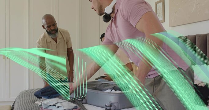 Packing man in pink polo leaning over suitcase in bedroom, with white headphones and tablet