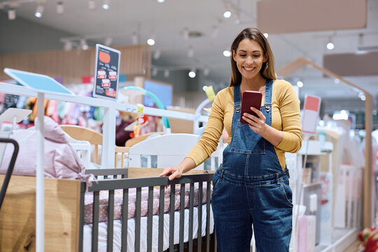 Happy pregnant woman using cell phone while buying baby crib at kids store. - Powered by Adobe