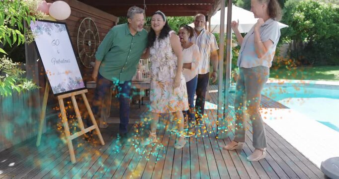 Woman walking to easel, host pointing, causing confetti sweeping group, guests hugging marking 60th - Powered by Adobe