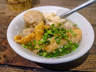 Close up of a bowl of bakso, an indonesian meatball soup, featuring meatballs, fried tofu, wontons, and garnished with fresh green onions, served on a wooden table, ready to eat.
