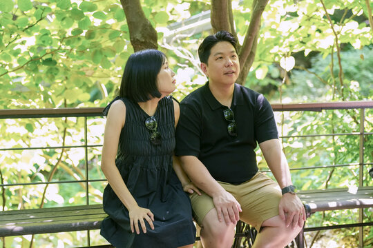 A man and woman are sitting on a bench in a park. The man is wearing sunglasses and the woman is wearing a black dress. Scene is relaxed and casual